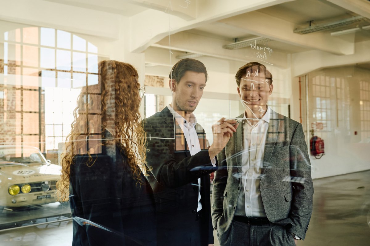 Drei Personen bei einer Besprechung, schreiben auf eine Glaswand. Industrieller Raum mit Oldtimer im Hintergrund