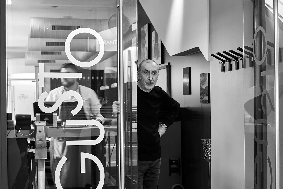 Schwarz-Weiss-Porträt Wirt an der Glastür des Restaurant. Auf der Tür in großen weißen Buchstaben: „Gusto“