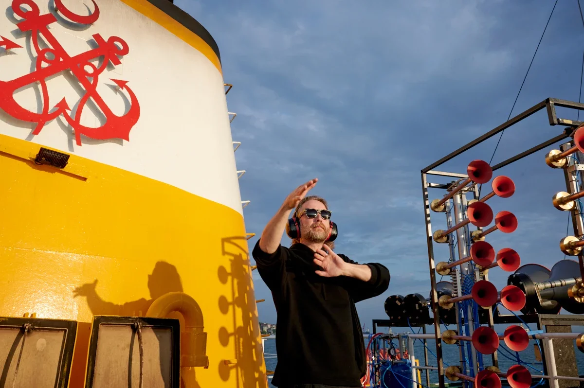 Der Tonkünstler Christoph Schläger auf einem Schiff neben einer Installtion mit Nebelhörnern. Während er dirigiert, trägt der Künstler Ohrenschützer. Reportagefoto von Nathalie Zimmermann