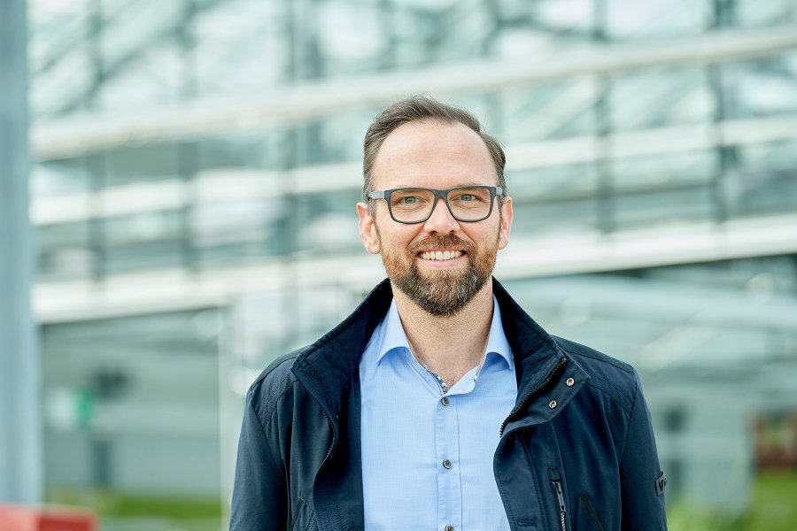 Mann mittleren alters  in Casual Kleidung blickt direkt in die Kamera, im Hintergrund unscharfe Fenster, freundliche Stimmung