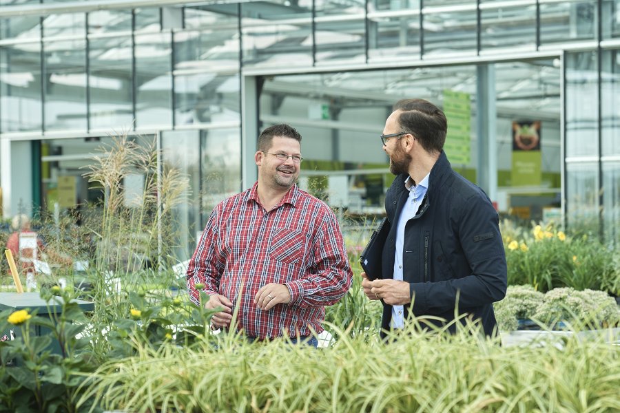 Gärtner im karierten Hemd in einer Gärtnerei am Gehen im Gespräch mit einem Mann
