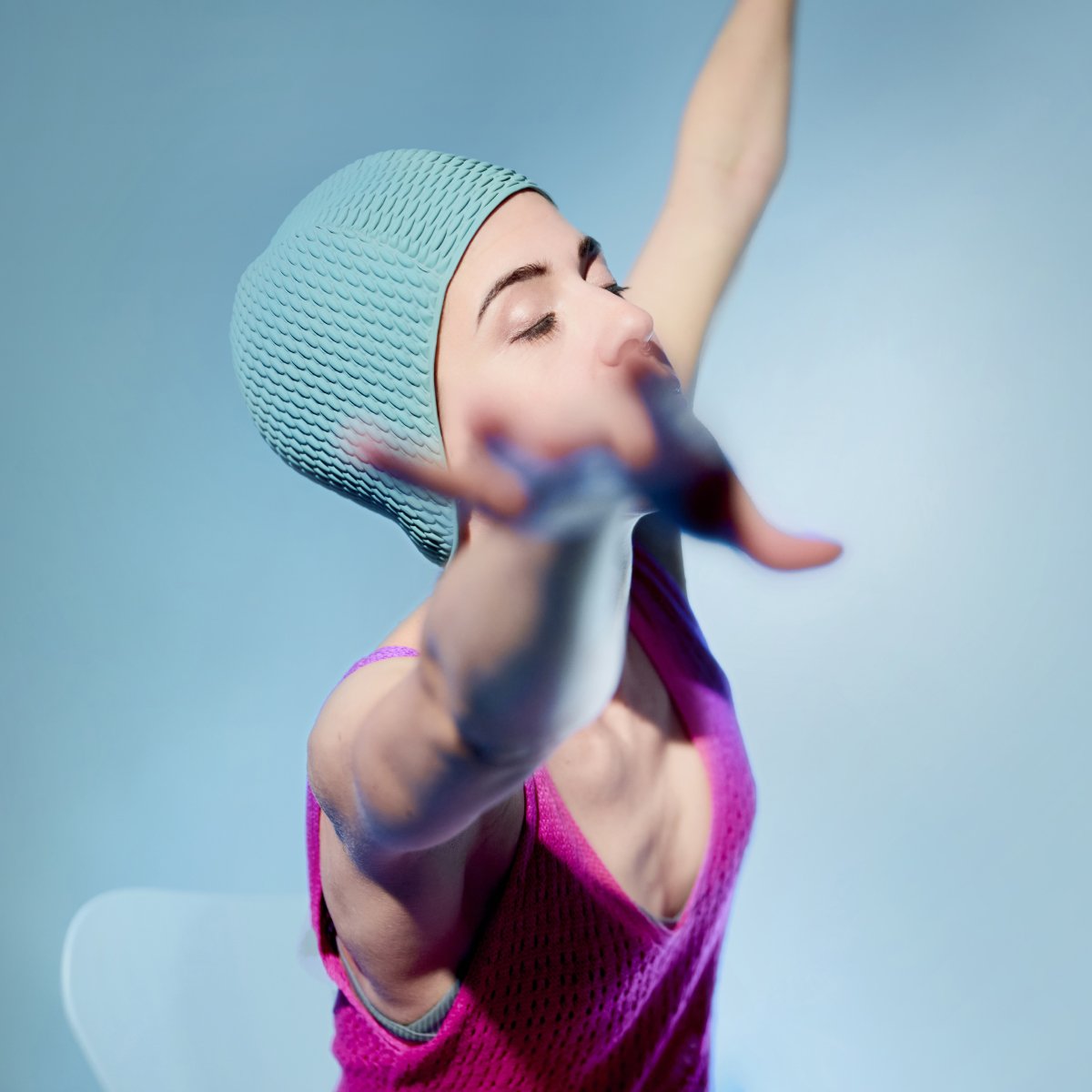 Frau mit Mint-farbener Badekappe und pinkem Strick-Kleid vor Mint-farbenem Studio-Hintergrund. Sie sitzt auf einem Stuhl und reckt mit geschlossenen Augen die Hände in die Luft.