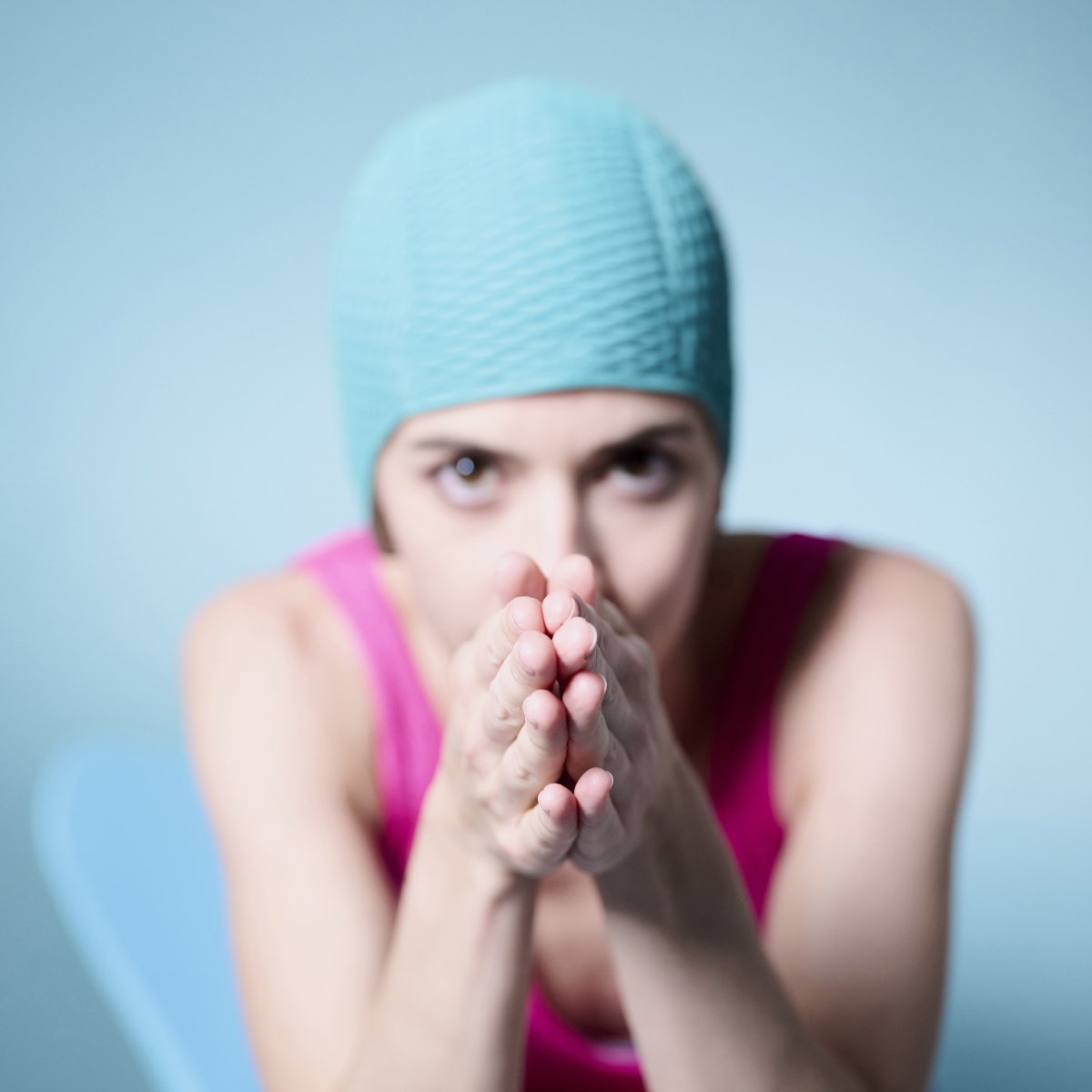 Frau mit Mint-farbener Badekappe vor Mint-farbenem Studio-Hintergrund. Sie hat die Hände wie beim Brustschwimmen zusammengelegt und blickt frontal in die Kamera.
