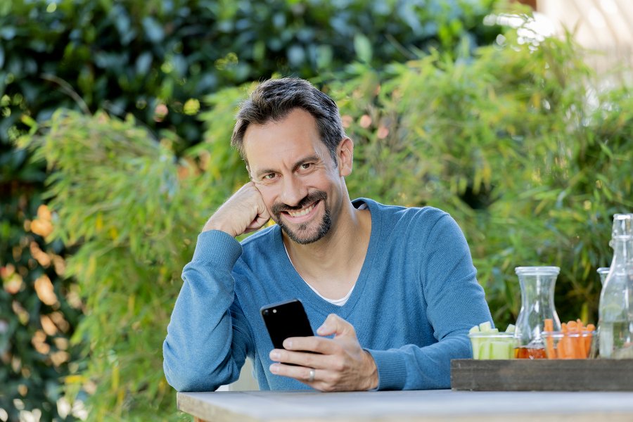 Mann mit dunkelbraunen Haaren sitzt im Garten mit Handy schaut in die Kamera nd lächelt.