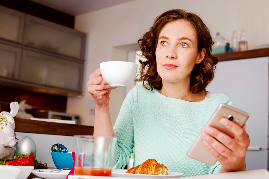 Frau mit Kaffetasse und Handy schaut nachdenklich.Woman with coffee cup and mobile phone looks thoughtful